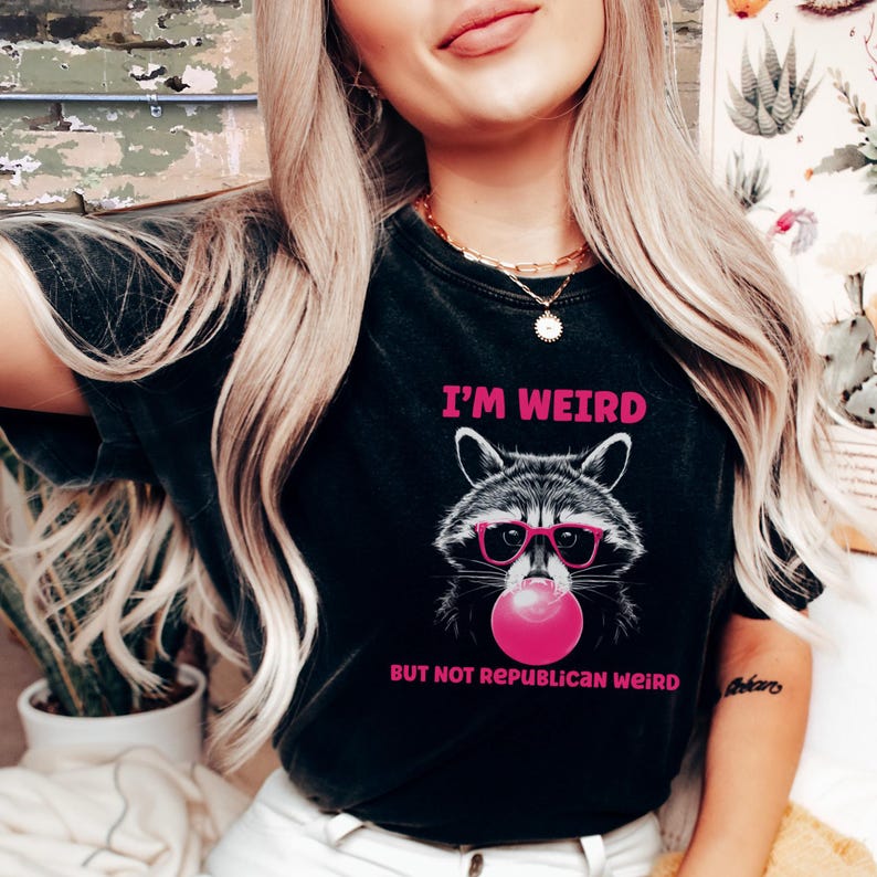 Peut inclure: T-shirt noir avec un raton laveur rose portant des lunettes de soleil roses et soufflant une bulle rose. Le t-shirt porte l'inscription "I'm Weird But Not Republican Weird".