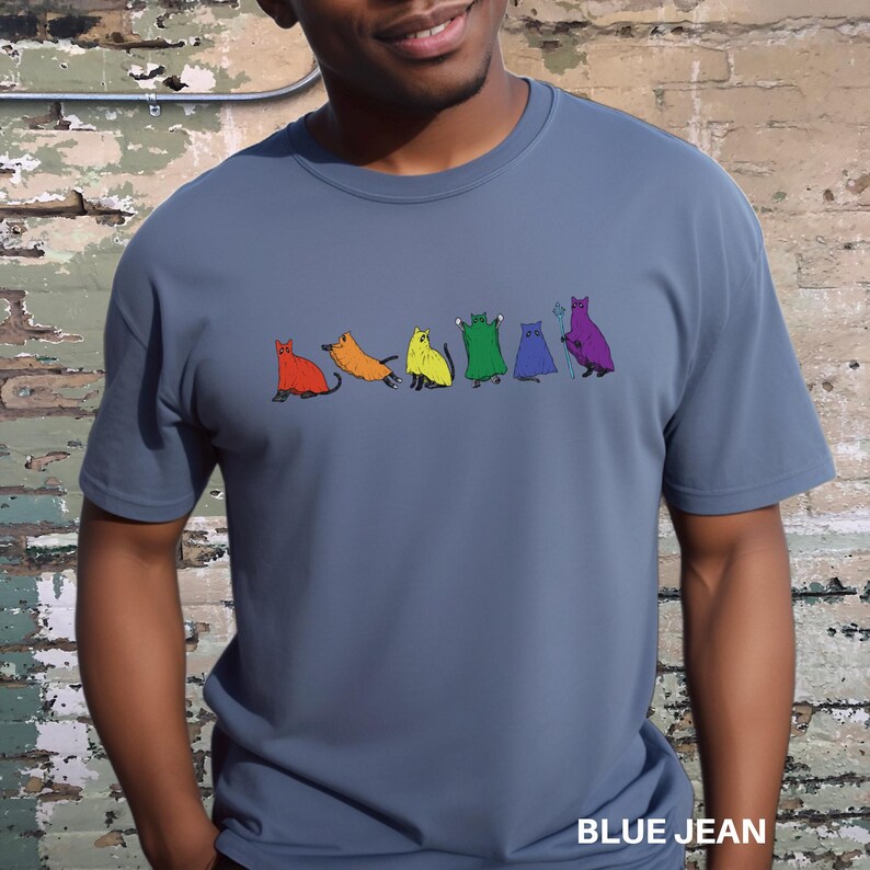 Peut inclure: T-shirt bleu jean avec une rang&eacute;e d'illustrations de chats color&eacute;s. Chaque chat est d'une couleur diff&eacute;rente, notamment rouge, orange, jaune, vert, bleu et violet. Le texte "BLUE JEAN" est imprim&eacute; en bas du t-shirt.
