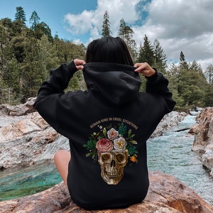 Puede incluir: Sudadera con capucha negra con un dise&ntilde;o de calavera y flores. El texto "REMAIN KIND IN CRUEL SITUATIONS" est&aacute; impreso en la parte posterior de la sudadera.