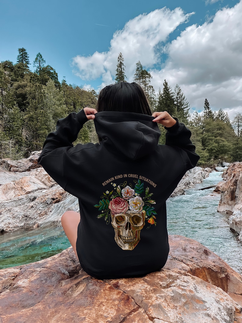 Puede incluir: Sudadera con capucha negra con un dise&ntilde;o de calavera y flores. El texto "REMAIN KIND IN CRUEL SITUATIONS" est&aacute; impreso en la parte posterior de la sudadera.