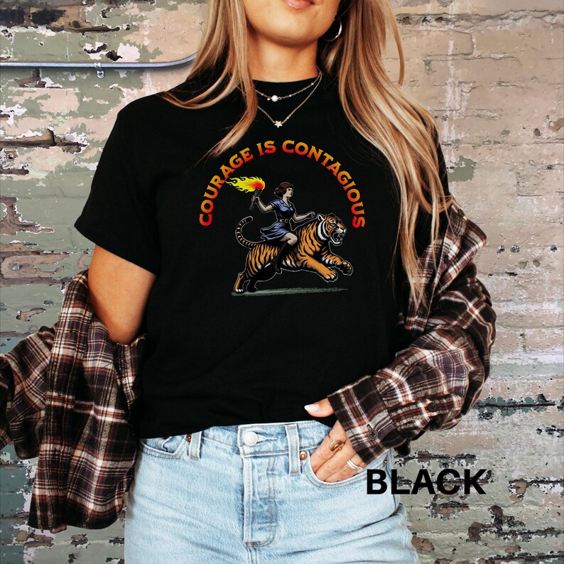 K&ouml;nnte beinhalten: Schwarzes T-Shirt mit einem Grafikdruck einer Frau, die auf einem Tiger reitet, mit dem Text "Courage is Contagious".