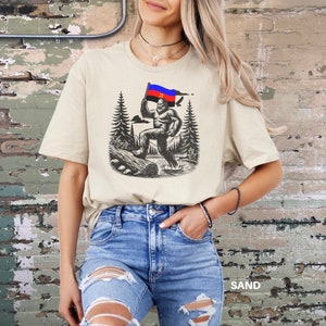 Peut inclure: T-shirt couleur sable avec une illustration en noir et blanc de Bigfoot tenant un drapeau &agrave; rayures rouges, bleues et noires. Le motif comprend des arbres et une b&ucirc;che. Le mot "SAND" est imprim&eacute; en bas.