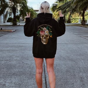Puede incluir: Sudadera negra con un dise&ntilde;o de calavera y flores en la espalda. El texto "REMAIN KIND IN CRUEL SITUATIONS" est&aacute; impreso en la sudadera.