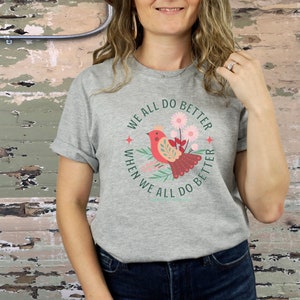 May include: Gray t-shirt with a red bird and green leaves in a circle. The text reads "We all do better when we all do better".