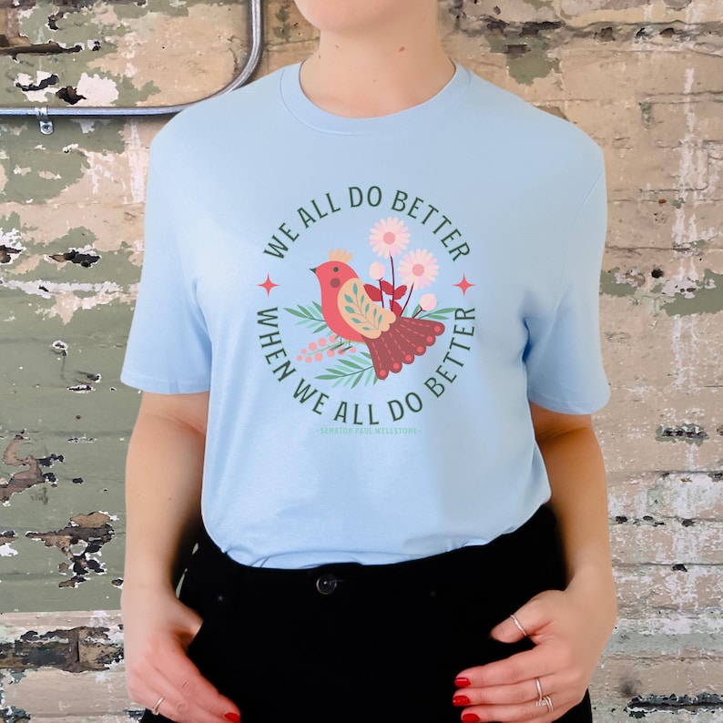 May include: Light blue t-shirt with a red bird and green floral design. The shirt has the text "We all do better when we all do better" in a circle around the bird.