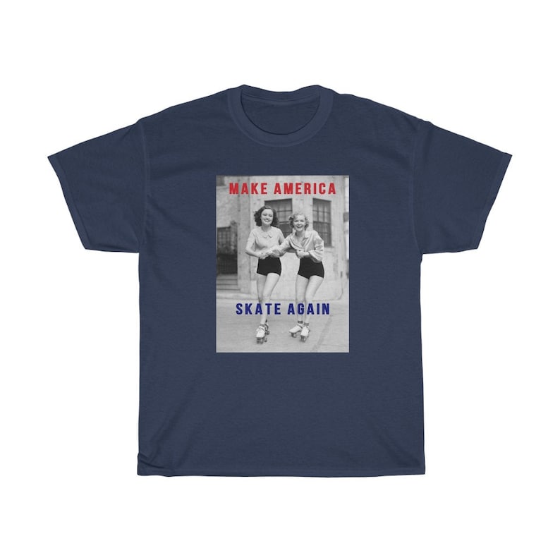 May include: Navy blue t-shirt featuring a graphic with two women roller skating. The graphic includes the text "MAKE AMERICA" in red and "SKATE AGAIN" in blue. The image is a vintage black and white photograph.