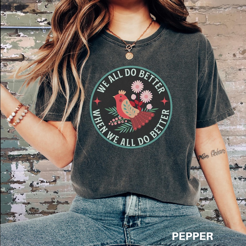 Puede incluir: Una camiseta gris oscuro con un gr&aacute;fico circular que presenta un p&aacute;jaro rojo con una corona, rodeado de flores y hojas. El texto "We all do better when we all do better" est&aacute; escrito alrededor del gr&aacute;fico.