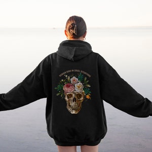Puede incluir: Sudadera con capucha negra con un cr&aacute;neo y flores bordadas en la espalda. El texto "Mantente amable en situaciones crueles" est&aacute; por encima del cr&aacute;neo.