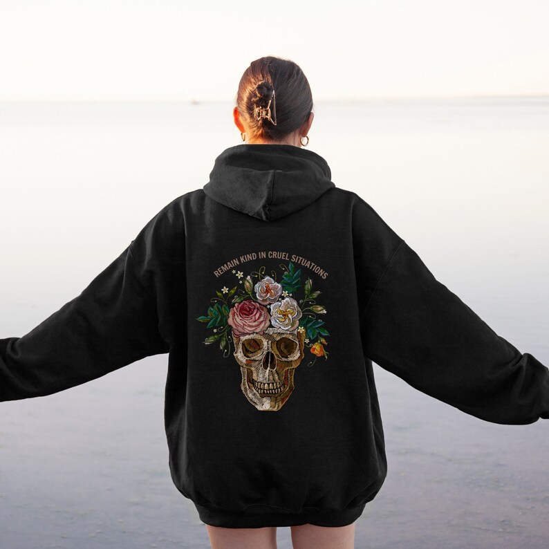 Puede incluir: Sudadera con capucha negra con un cr&aacute;neo y flores bordadas en la espalda. El texto "Mantente amable en situaciones crueles" est&aacute; por encima del cr&aacute;neo.