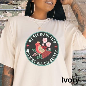 Puede incluir: Una camiseta blanca con un gr&aacute;fico de un p&aacute;jaro y flores en un c&iacute;rculo. El texto "We all do better when we all do better" est&aacute; escrito alrededor del c&iacute;rculo. La camiseta est&aacute; disponible en marfil.