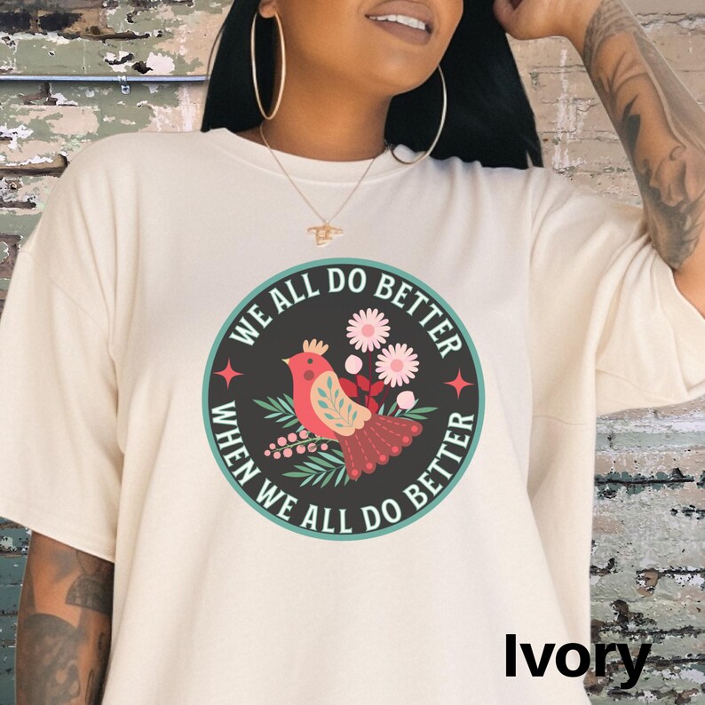 Puede incluir: Una camiseta blanca con un gr&aacute;fico de un p&aacute;jaro y flores en un c&iacute;rculo. El texto "We all do better when we all do better" est&aacute; escrito alrededor del c&iacute;rculo. La camiseta est&aacute; disponible en marfil.