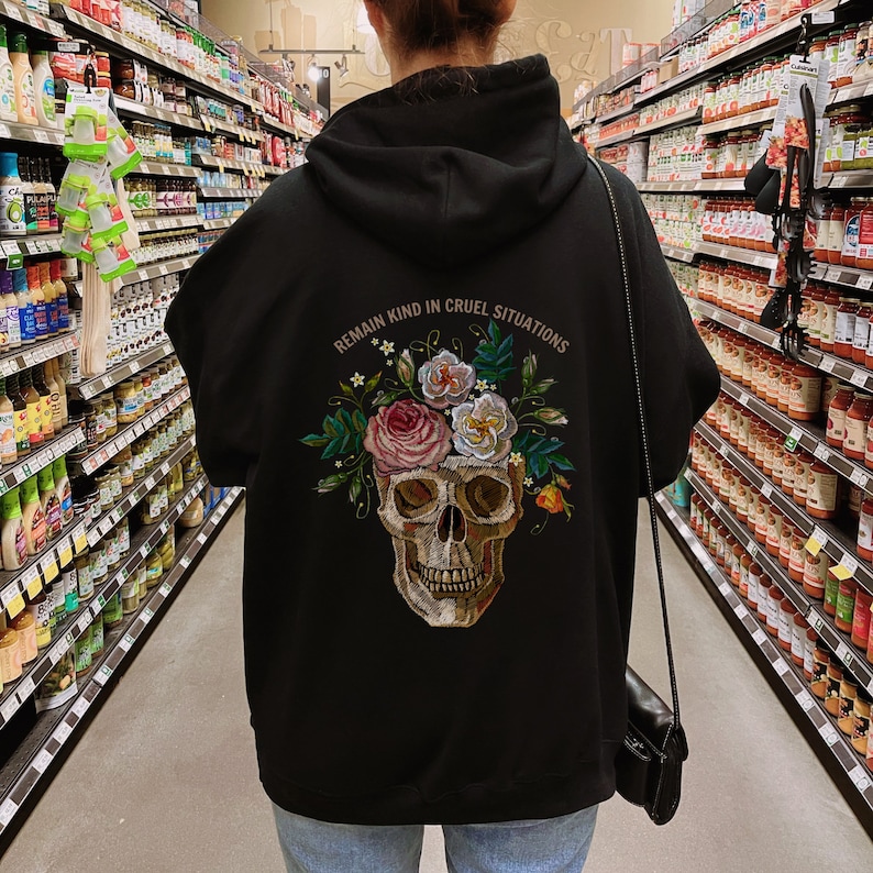 Puede incluir: Sudadera con capucha negra con un dise&ntilde;o de calavera y flores. El texto "Mantente amable en situaciones crueles" est&aacute; impreso en la parte posterior de la sudadera.