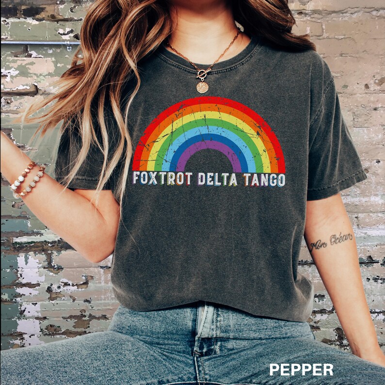 May include: A dark gray t-shirt with a rainbow graphic and the text "FOXTROT DELTA TANGO" printed on the front. The shirt is made of a soft, comfortable material and has a relaxed fit.