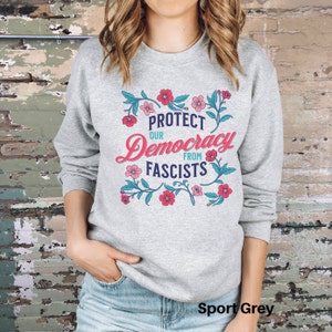 May include: A gray sweatshirt with a floral design and the text "Protect Our Democracy From Fascists". The sweatshirt is a sport grey color.