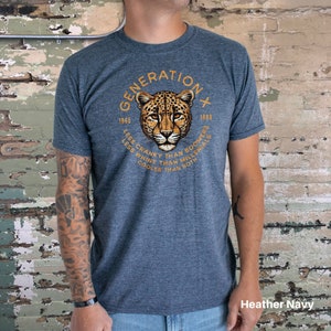 Puede incluir: Una camiseta azul marino jaspeada con un gr&aacute;fico de la cara de un guepardo en un c&iacute;rculo. El c&iacute;rculo tiene el texto "GENERATION X" en la parte superior, "1965" y "1980" en la parte inferior, y el texto "LESS CRANKY THAN BOOMERS, LESS WHINY THAN MILLENIALS, COOLER THAN BOTH" alrededor de la cara del guepardo.