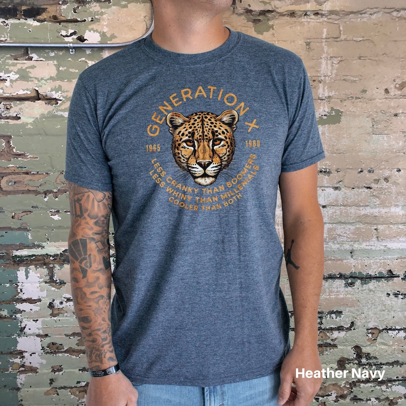 Puede incluir: Una camiseta azul marino jaspeada con un gr&aacute;fico de la cara de un guepardo en un c&iacute;rculo. El c&iacute;rculo tiene el texto "GENERATION X" en la parte superior, "1965" y "1980" en la parte inferior, y el texto "LESS CRANKY THAN BOOMERS, LESS WHINY THAN MILLENIALS, COOLER THAN BOTH" alrededor de la cara del guepardo.