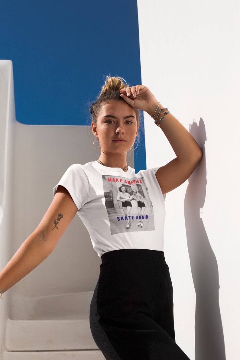 May include: White t-shirt with the text "MAKE AMERICA SKATE AGAIN" above a black and white image of two women roller skating. The model is wearing black pants and has a small tattoo on her arm.