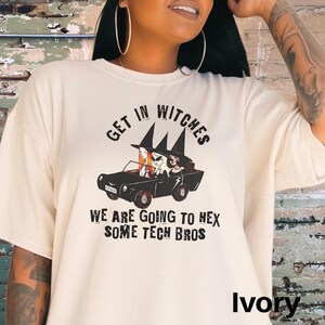 Może przedstawiać: Biały t-shirt z czarnym nadrukiem przedstawiającym trzy czarownice prowadzące samoch&oacute;d. Tekst na koszulce brzmi "Get in Witches We are going to hex some tech bros".