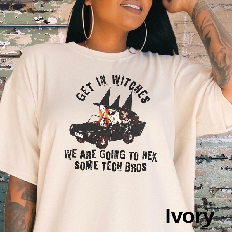Może przedstawiać: Biały t-shirt z czarnym nadrukiem przedstawiającym trzy czarownice prowadzące samoch&oacute;d. Tekst na koszulce brzmi "Get in Witches We are going to hex some tech bros".