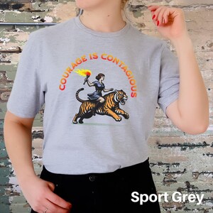 K&ouml;nnte beinhalten: Ein graues T-Shirt mit dem Text "Courage is Contagious" in einem kreisf&ouml;rmigen Design. Eine Frau, die auf einem Tiger mit einer Fackel reitet, ist in der Mitte des Kreises abgebildet. Der Text "Sport Grey" ist am unteren Rand des Bildes sichtbar.