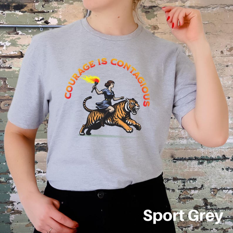 K&ouml;nnte beinhalten: Ein graues T-Shirt mit dem Text "Courage is Contagious" in einem kreisf&ouml;rmigen Design. Eine Frau, die auf einem Tiger mit einer Fackel reitet, ist in der Mitte des Kreises abgebildet. Der Text "Sport Grey" ist am unteren Rand des Bildes sichtbar.
