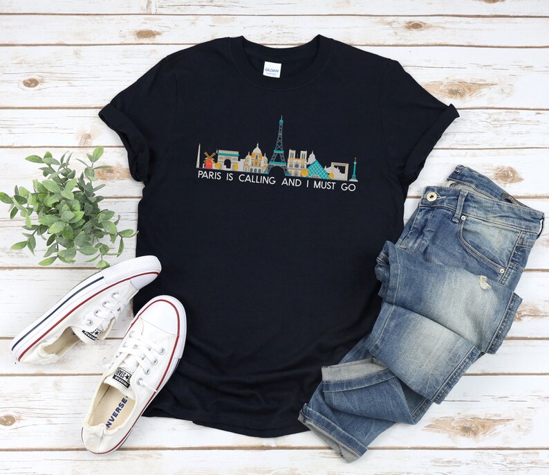 May include: A navy blue t-shirt with a graphic of the Paris skyline and the text "Paris is calling and I must go". The t-shirt is laid out on a wooden surface with a pair of white sneakers and blue jeans.
