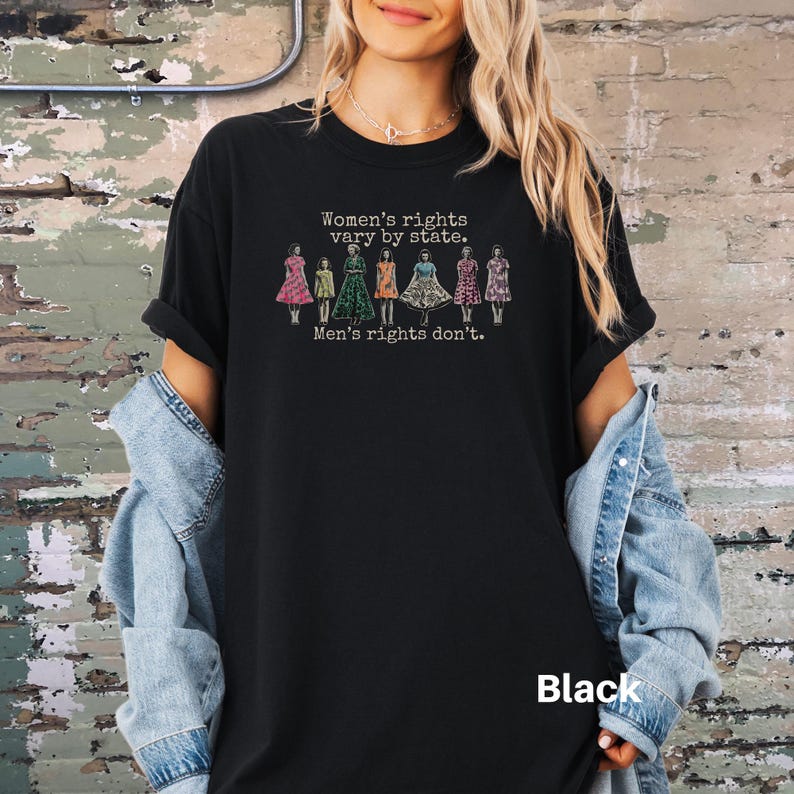 Puede incluir: Camiseta negra con el texto "Women's rights vary by state. Men's rights don't." y un gr&aacute;fico de mujeres con vestidos. La palabra "Black" est&aacute; impresa en la parte inferior derecha de la camiseta.