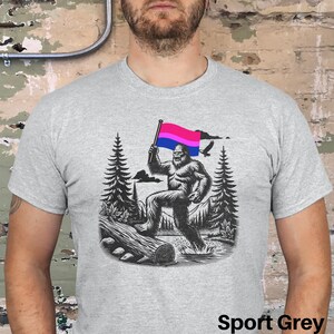 May include: A grey t-shirt with a graphic of a Bigfoot holding a bisexual pride flag in a forest setting. The text "Sport Grey" is visible at the bottom of the image.