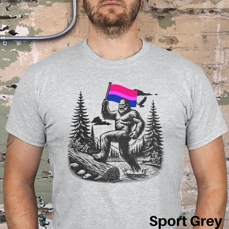 May include: A grey t-shirt with a graphic of a Bigfoot holding a bisexual pride flag in a forest setting. The text "Sport Grey" is visible at the bottom of the image.