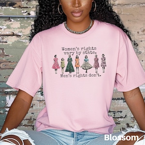 Puede incluir: Una camiseta rosa claro con el texto "Women's rights vary by state. Men's rights don't." sobre un gr&aacute;fico de siete mujeres con vestidos. La palabra "Blossom" est&aacute; impresa en blanco en la parte inferior derecha.