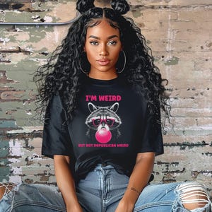 Peut inclure: Un t-shirt noir avec un graphique d'un raton laveur portant des lunettes de soleil et soufflant une bulle rose. Le texte sur le t-shirt est "I'm Weird But Not Republican Weird."