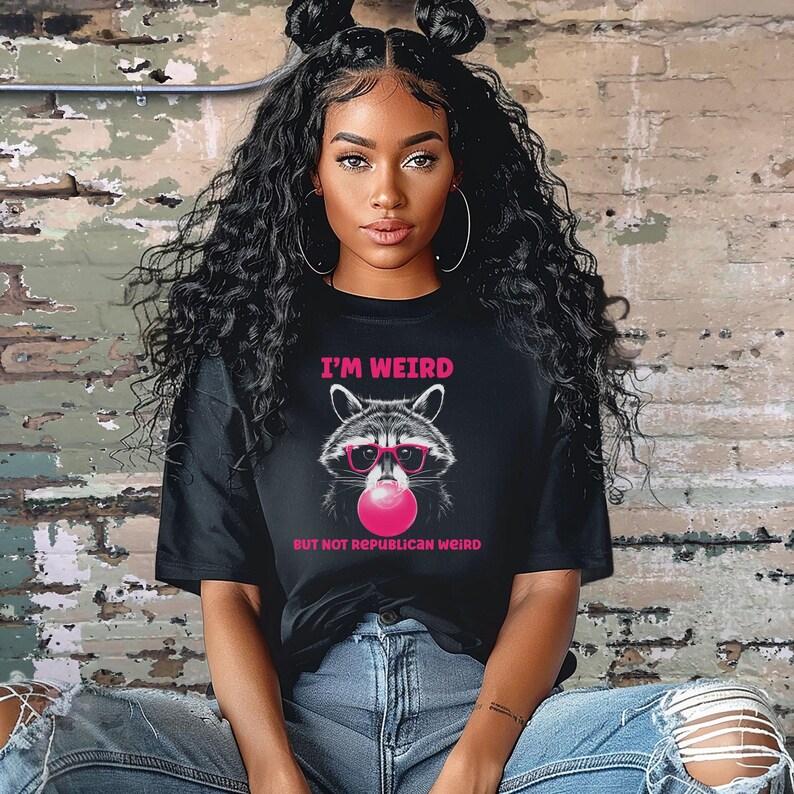 Peut inclure: Un t-shirt noir avec un graphique d'un raton laveur portant des lunettes de soleil et soufflant une bulle rose. Le texte sur le t-shirt est "I'm Weird But Not Republican Weird."