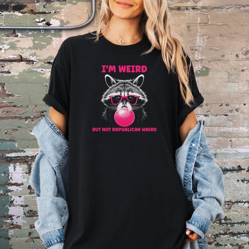 Peut inclure: T-shirt noir avec un graphique d'un raton laveur portant des lunettes de soleil et soufflant une bulle rose. Le texte "I'm Weird But Not Republican Weird" est imprim&eacute; sur le t-shirt.