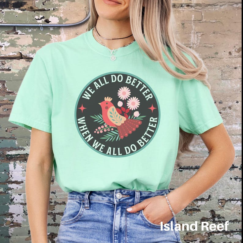 Puede incluir: Una camiseta verde claro con un gr&aacute;fico redondo que presenta un p&aacute;jaro rojo con una corona, flores y el texto "We all do better when we all do better".