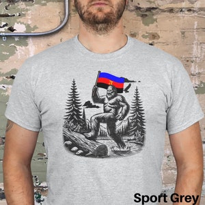 Peut inclure: T-shirt gris sport avec une illustration en noir et blanc d'un Bigfoot tenant un drapeau aux couleurs rouge, bleu et noir. Le drapeau porte les initiales "JT". Le motif comprend des arbres, une b&ucirc;che et un oiseau en vol.