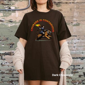 K&ouml;nnte beinhalten: Ein dunkelbraunes T-Shirt mit dem Text "Courage is Contagious" in einem Kreis um eine Frau, die auf einem Tiger reitet. Die Frau h&auml;lt eine Fackel.