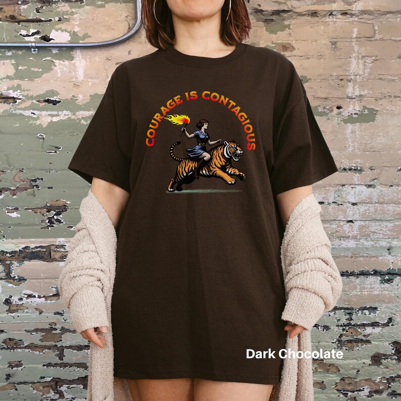 K&ouml;nnte beinhalten: Ein dunkelbraunes T-Shirt mit dem Text "Courage is Contagious" in einem Kreis um eine Frau, die auf einem Tiger reitet. Die Frau h&auml;lt eine Fackel.