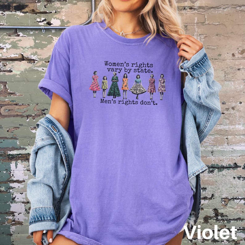 Puede incluir: Una camiseta de color violeta con el texto "Women's rights vary by state. Men's rights don't." impreso en negro. La camiseta presenta una l&iacute;nea de figuras ilustradas con varios vestidos. La palabra "Violet" est&aacute; impresa en la parte inferior.