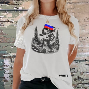 Peut inclure: T-shirt blanc avec une illustration en noir et blanc d'un Bigfoot tenant un drapeau aux couleurs rouge, bleu et jaune. Le motif comprend des arbres, une b&ucirc;che et un oiseau. Le mot "WHITE" est imprim&eacute; en bas.