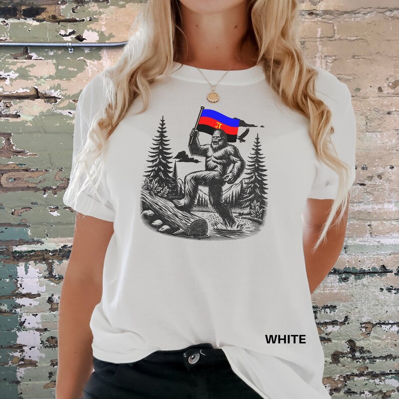 Peut inclure: T-shirt blanc avec une illustration en noir et blanc d'un Bigfoot tenant un drapeau aux couleurs rouge, bleu et jaune. Le motif comprend des arbres, une b&ucirc;che et un oiseau. Le mot "WHITE" est imprim&eacute; en bas.