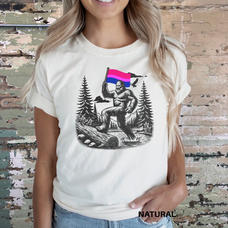 May include: A white t-shirt with a graphic of a Bigfoot holding a bisexual pride flag in a forest setting.
