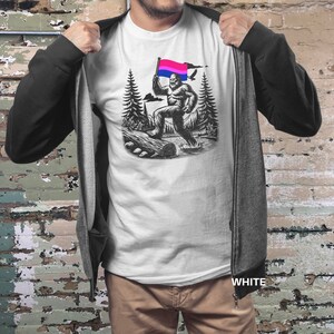 May include: A white t-shirt with a black and white illustration of a Bigfoot holding a bisexual pride flag. The Bigfoot is walking through a forest with trees and a log in the background.