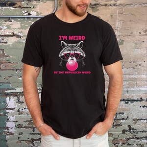 Peut inclure: T-shirt noir avec un graphique d'un raton laveur portant des lunettes de soleil roses et soufflant une bulle rose. Le texte "I'm Weird But Not Republican Weird" est imprim&eacute; sous le raton laveur.