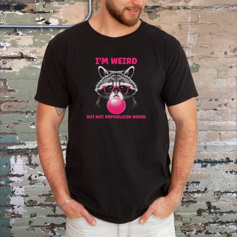 Peut inclure: T-shirt noir avec un graphique d'un raton laveur portant des lunettes de soleil roses et soufflant une bulle rose. Le texte "I'm Weird But Not Republican Weird" est imprim&eacute; sous le raton laveur.