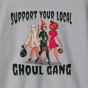 K&ouml;nnte beinhalten: Ein graumeliertes T-Shirt mit einer Grafik von drei Figuren in Halloween-Kost&uuml;men: ein Geist, ein Skelett und eine Person mit K&uuml;rbiskopf. Der Text "SUPPORT YOUR LOCAL GHOUL GANG" ist in Schwarz mit gr&uuml;nem Umriss &uuml;ber und unter den Figuren gedruckt.