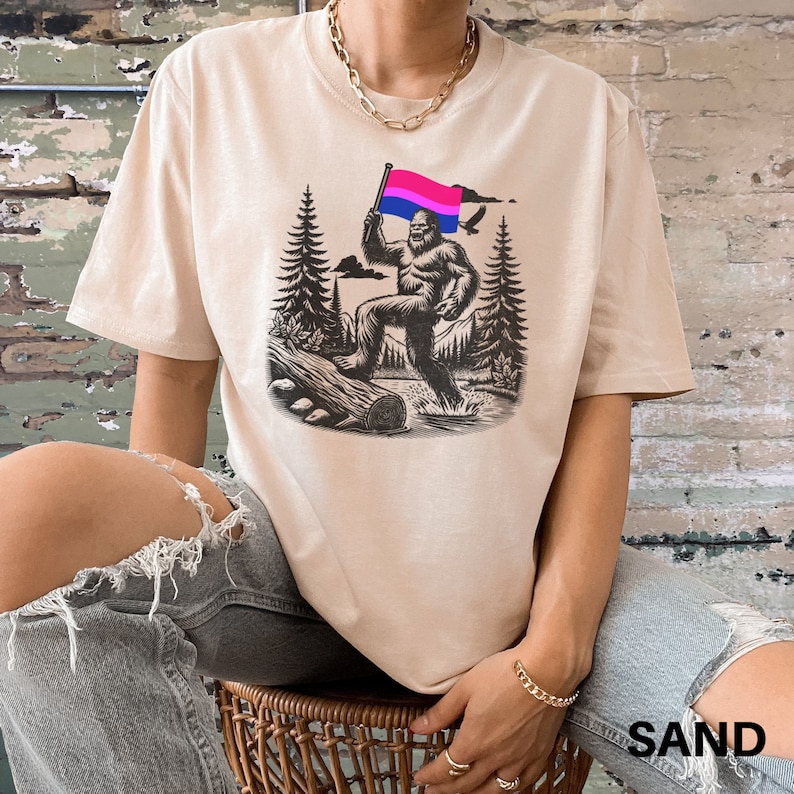 May include: A beige t-shirt with a graphic print of a Bigfoot holding a bisexual pride flag. The Bigfoot is standing in a forest setting with trees and a log.