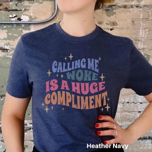 May include: A heather navy blue t-shirt with the text "Calling me woke is a huge compliment" in a retro font with colourful accents and gold stars.