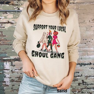K&ouml;nnte beinhalten: Ein sandfarbenes Sweatshirt mit Rundhalsausschnitt und einem Grafikdruck von drei Figuren in Halloween-Kost&uuml;men: ein Geist, ein Skelett und eine Figur mit K&uuml;rbiskopf. Der Text "SUPPORT YOUR LOCAL GHOUL GANG" ist in Schwarz &uuml;ber und unter den Figuren gedruckt.