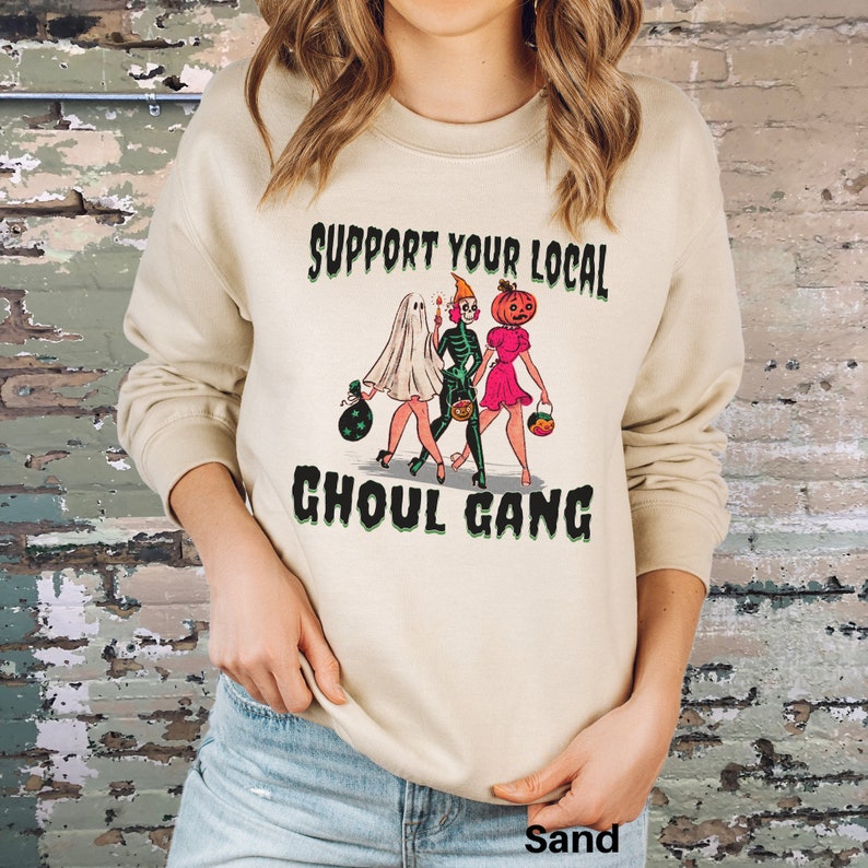 K&ouml;nnte beinhalten: Ein sandfarbenes Sweatshirt mit Rundhalsausschnitt und einem Grafikdruck von drei Figuren in Halloween-Kost&uuml;men: ein Geist, ein Skelett und eine Figur mit K&uuml;rbiskopf. Der Text "SUPPORT YOUR LOCAL GHOUL GANG" ist in Schwarz &uuml;ber und unter den Figuren gedruckt.
