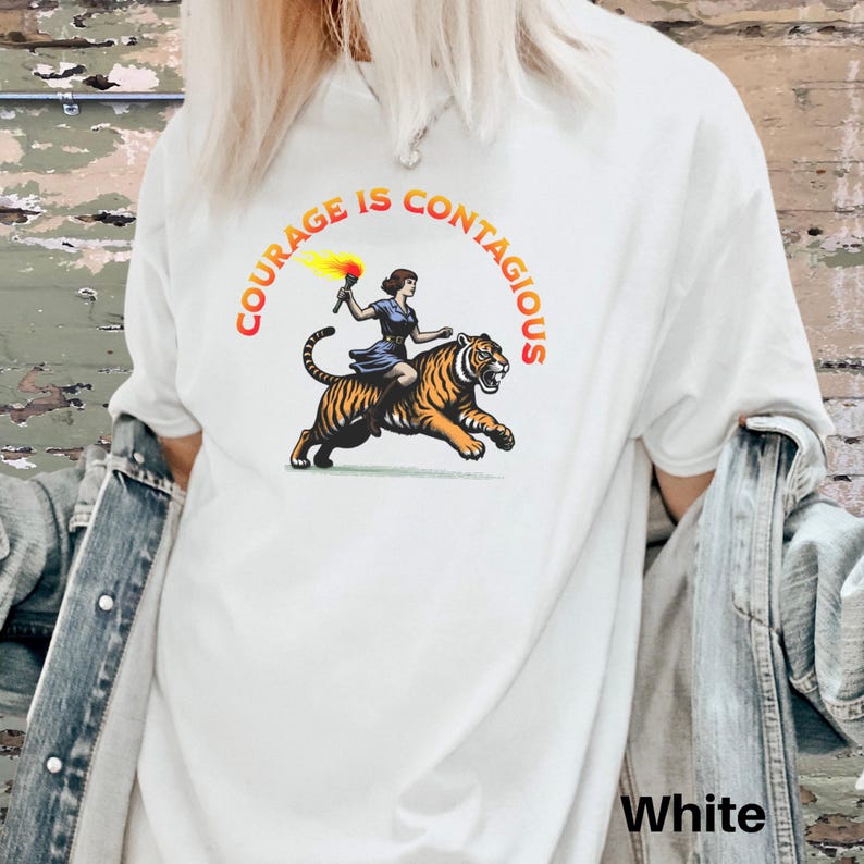 K&ouml;nnte beinhalten: Wei&szlig;es T-Shirt mit einem Motiv einer Frau, die auf einem Tiger mit einer Fackel reitet. Der Text "Courage is Contagious" ist in einem Bogen &uuml;ber dem Motiv gedruckt.